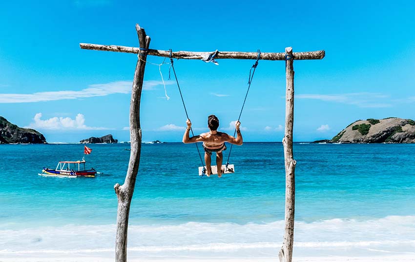 Young traveler guy is standing on swing on the beautiful beach in Bali. He is enjoying the Gloomy sand beach with the turquoise sea background at the paradise of indonesia. backpacker make a trip .